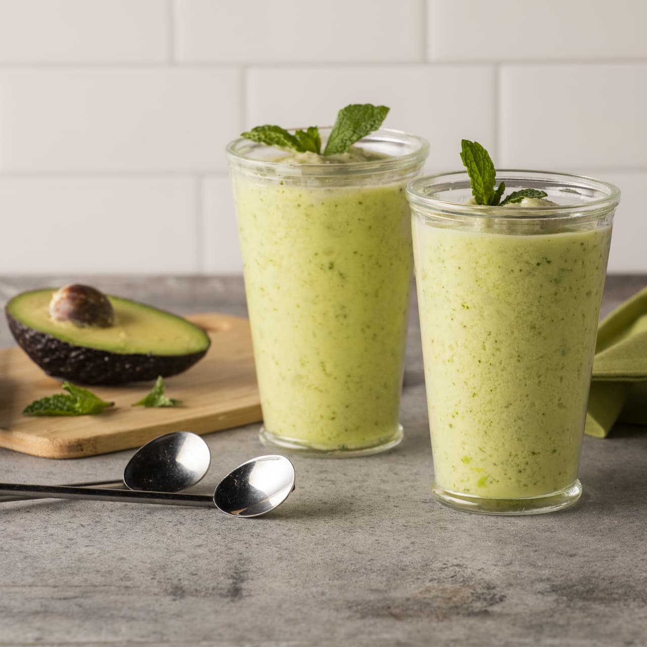 Two glasses of green smoothie garnished with mint, with two spoons and a halved avocado on a cutting board in the background.