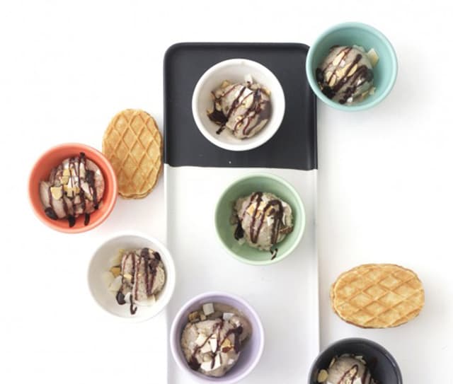 Six bowls of ice cream with chocolate drizzle and almonds are arranged on a white surface with waffle cookies placed nearby.