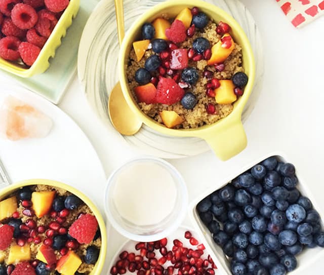 Bowls of oatmeal topped with blueberries, raspberries, pomegranate seeds, and peaches, surrounded by additional bowls of berries, pomegranate seeds, and a glass of milk.
