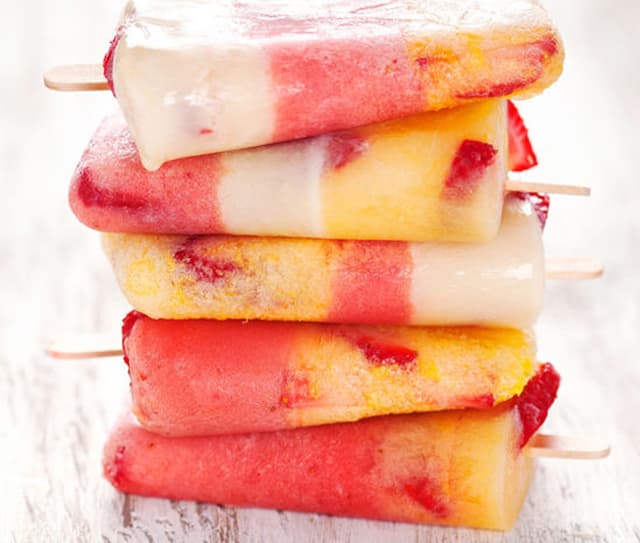 A stack of five layered fruit popsicles with red, yellow, and white sections, placed on a light wooden surface.