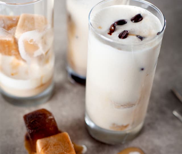 Three glasses of iced coffee with milk, some with coffee ice cubes. Whole coffee beans and ice cubes are visible in the drinks and on the gray tabletop.