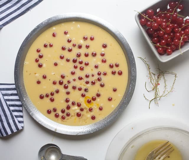 A round metal pan filled with yellow batter and scattered red currants, next to a small bowl of red currants, striped cloth, and utensils on a white surface.
