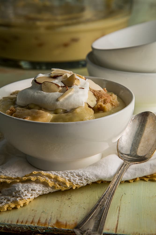 A bowl of pudding topped with whipped cream and sliced almonds sits on a napkin next to a spoon, with an empty bowl and more pudding in the background.