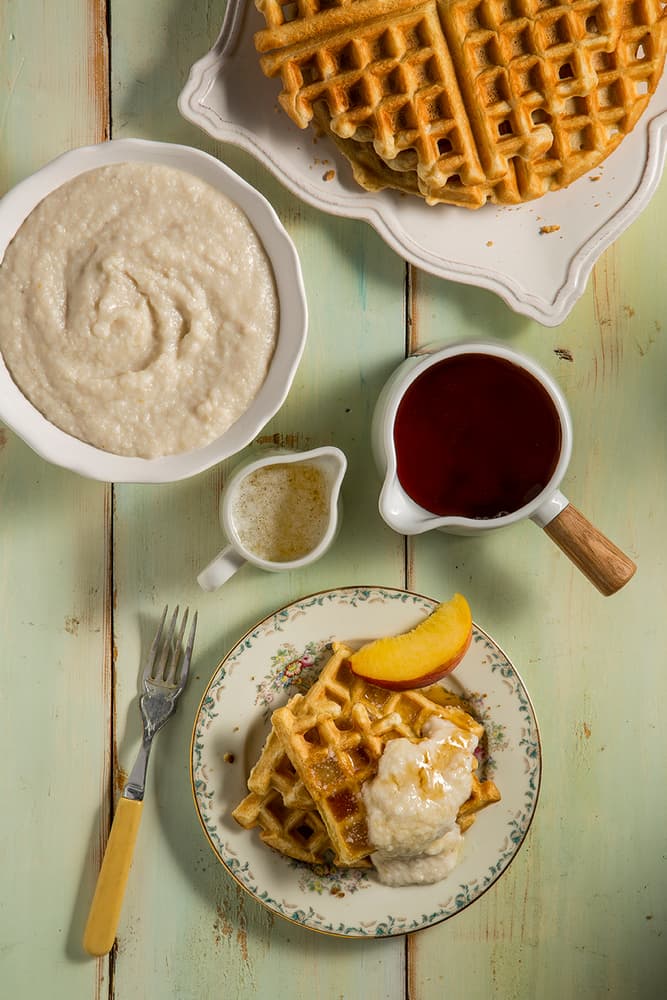 Plate with waffles, peach slice, and cream sauce. Nearby are a bowl of grits, stacked waffles, syrup pitcher, and small creamer on a rustic wooden table.