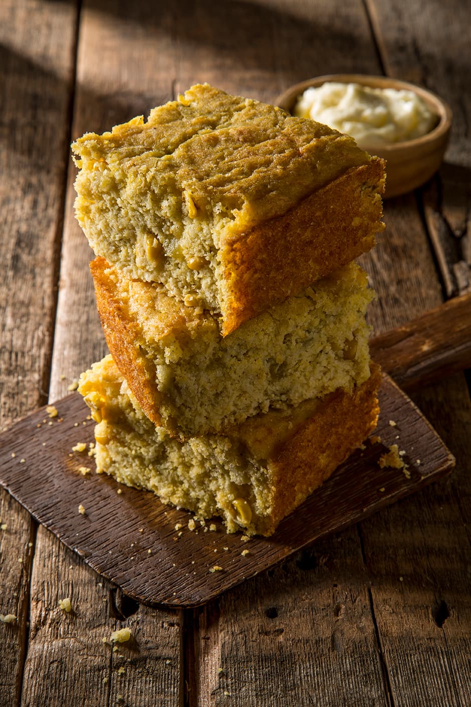 Three thick slices of cornbread are stacked on a wooden board, with a small bowl of butter or spread in the background, all placed on a rustic wooden table.
