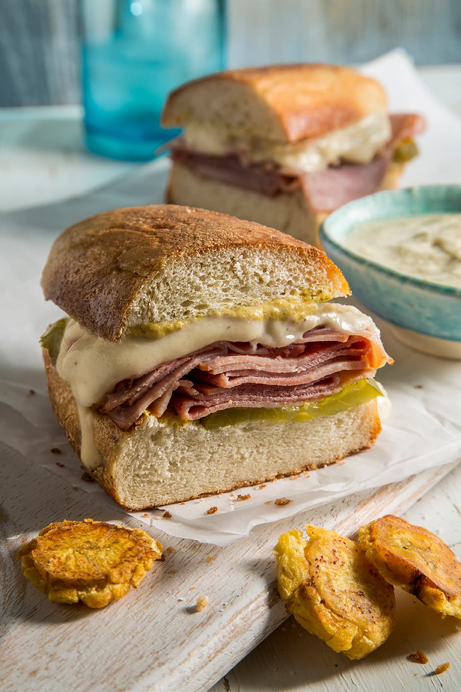 A close-up of a sandwich with ham, melted cheese, pickles, and sauce on thick bread, with fried plantain slices and a bowl of dip on the side.