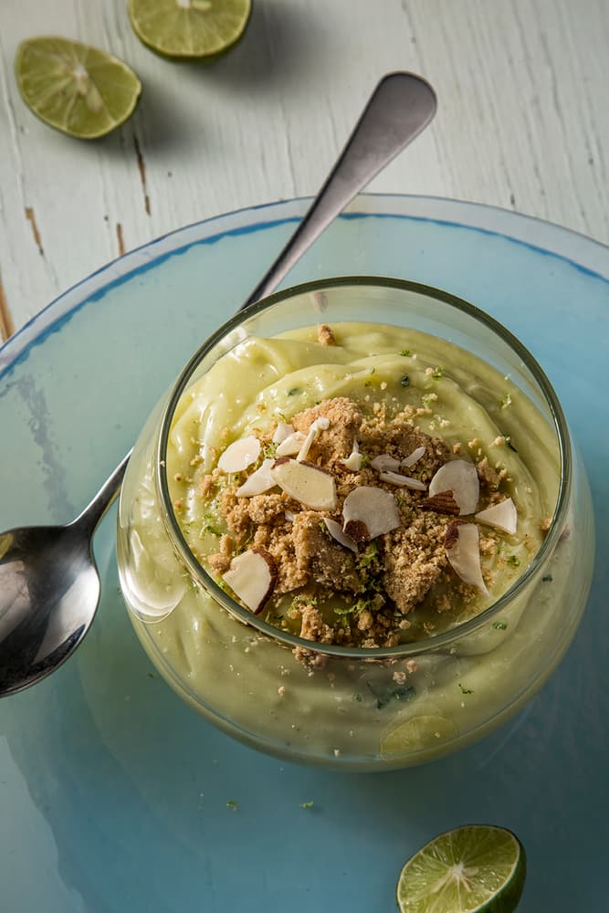 A glass bowl of creamy avocado dessert topped with cookie crumbs and almond slices, placed on a blue plate with a spoon and lime halves nearby.