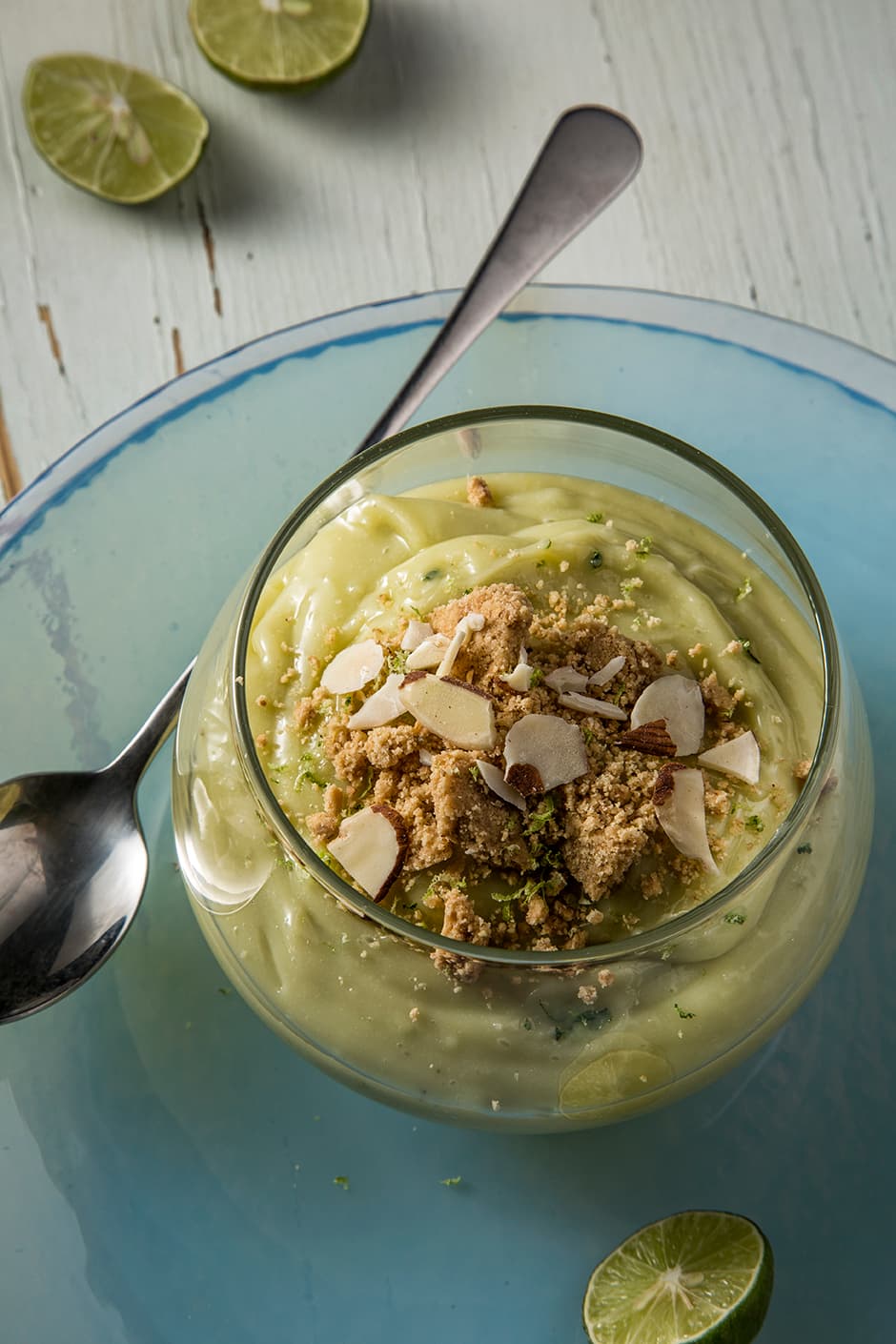 A glass bowl of creamy avocado dessert topped with cookie crumbs and almond slices, placed on a blue plate with a spoon and lime halves nearby.