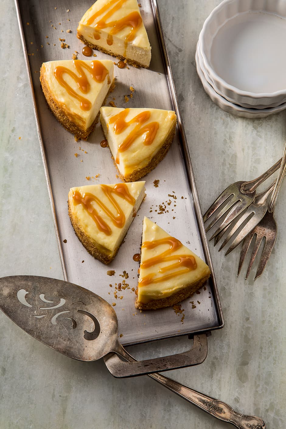 Four slices of cheesecake with caramel drizzle on a rectangular tray, with vintage serving utensils and two stacked white plates nearby.