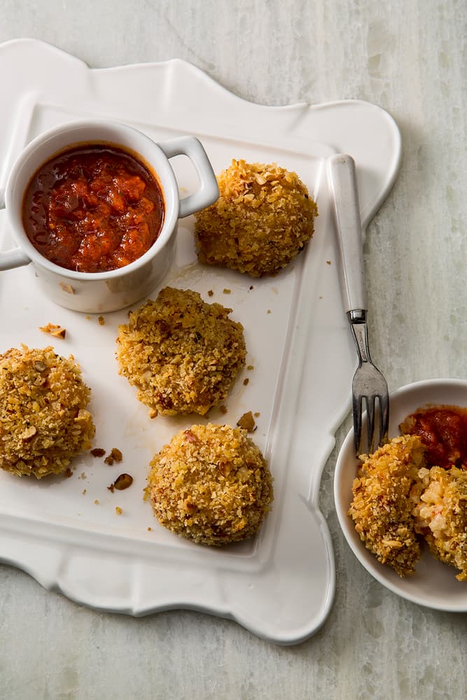 Five breaded, baked balls are arranged on a white tray with a small cup of chunky red dipping sauce; a fork and small bowl with sauce and one ball are nearby.