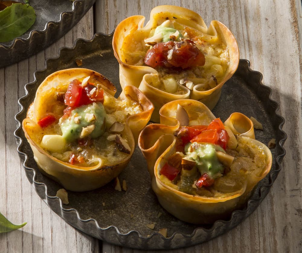 Three savory pastry cups filled with melted cheese, diced tomatoes, and green sauce are arranged in a round baking tin on a wooden surface.