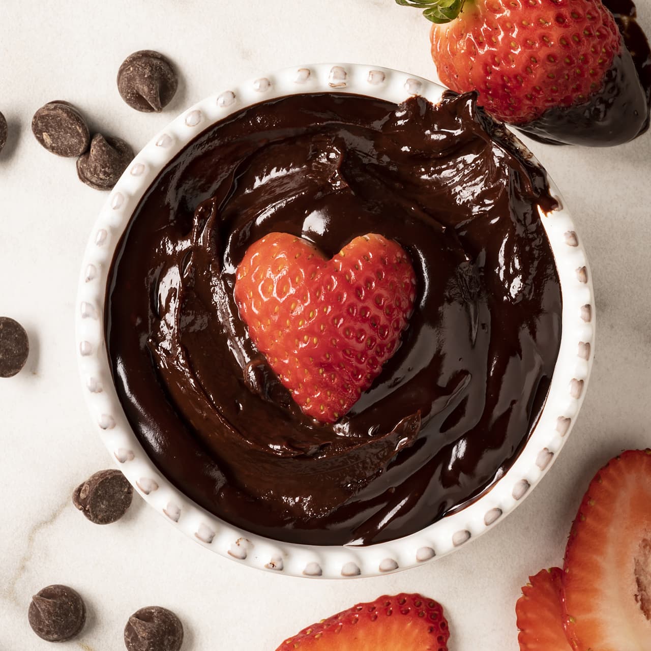 A heart-shaped strawberry sits in a bowl of chocolate sauce, surrounded by chocolate chips and sliced strawberries on a white surface.