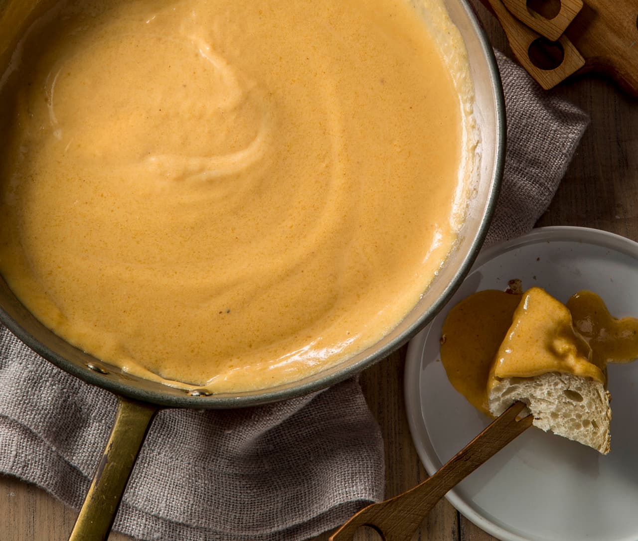 A saucepan filled with creamy cheese sauce sits on a cloth; a piece of bread with cheese sauce is on a small plate beside it.