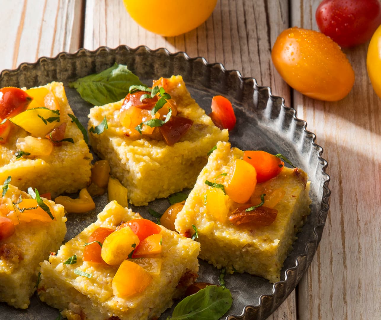Four square slices of cornbread topped with diced tomatoes and herbs are arranged on a metal plate, with whole tomatoes and basil leaves on the wooden table nearby.