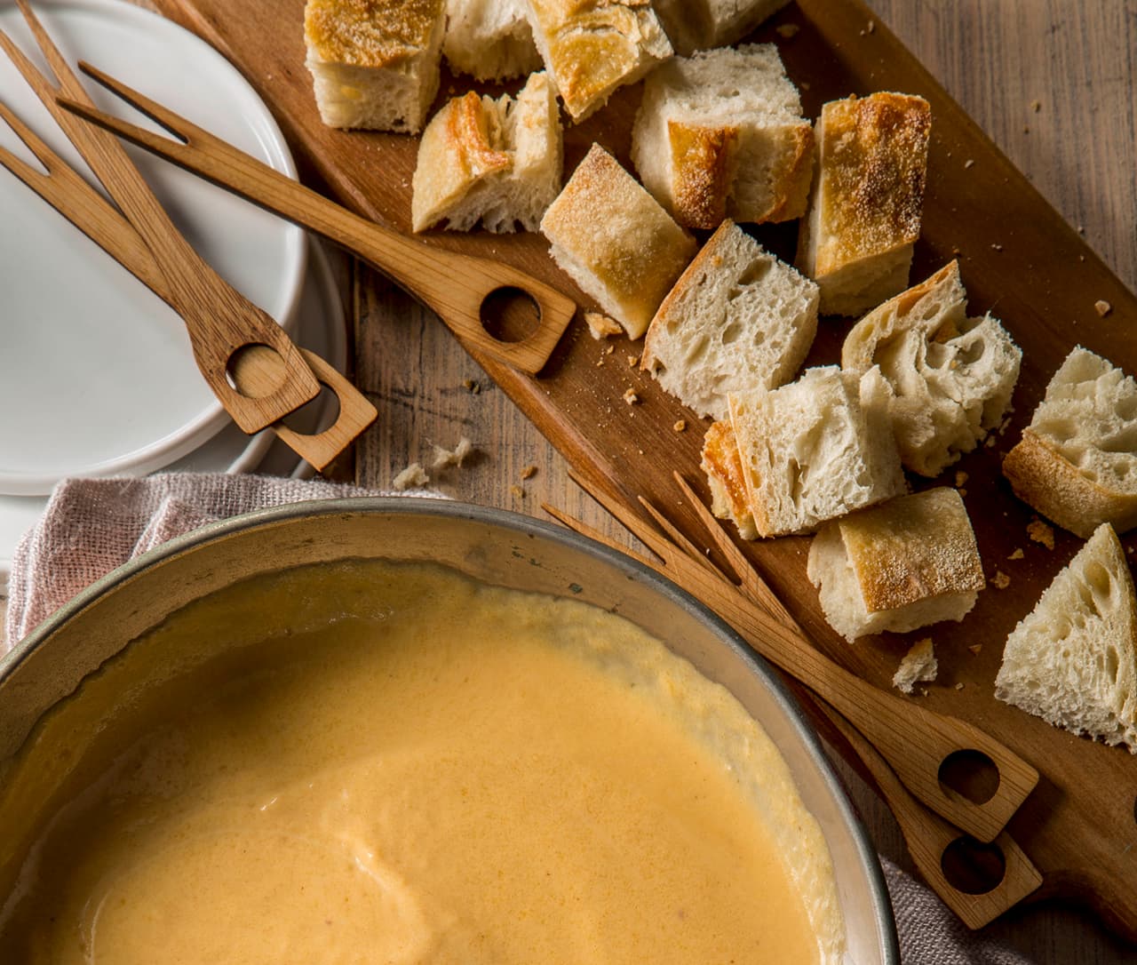 A pot of cheese fondue sits next to a wooden board topped with cubed bread and wooden fondue forks on a rustic table.