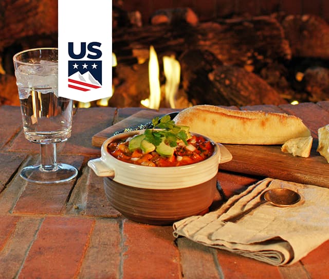 A bowl of chili topped with avocado and cilantro, a baguette, a glass of water, and a folded napkin are set on a brick surface near a fireplace.