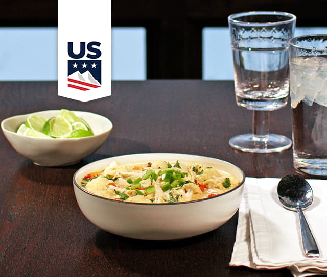 A bowl of creamy soup garnished with herbs sits on a table next to lime wedges, two glasses of water, a spoon, and a white napkin. A US logo appears at the top left.