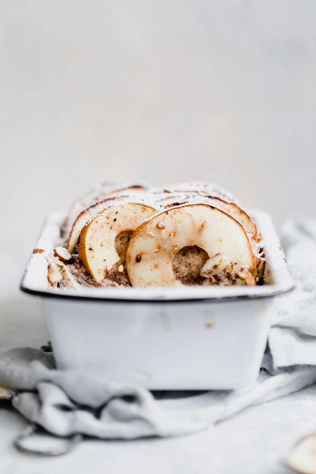 A white loaf pan filled with baked bread, topped with thinly sliced apples and a dusting of powdered sugar, sits on a light gray cloth.