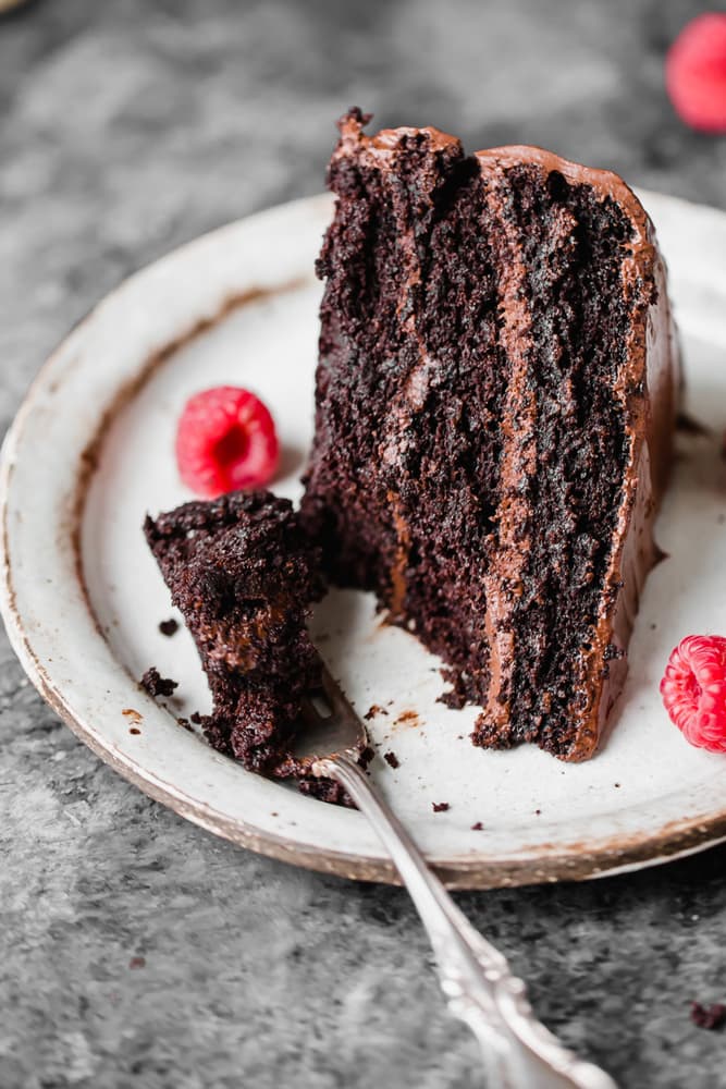 A slice of chocolate cake with chocolate frosting sits on a plate with raspberries; a fork holds a piece of the cake.