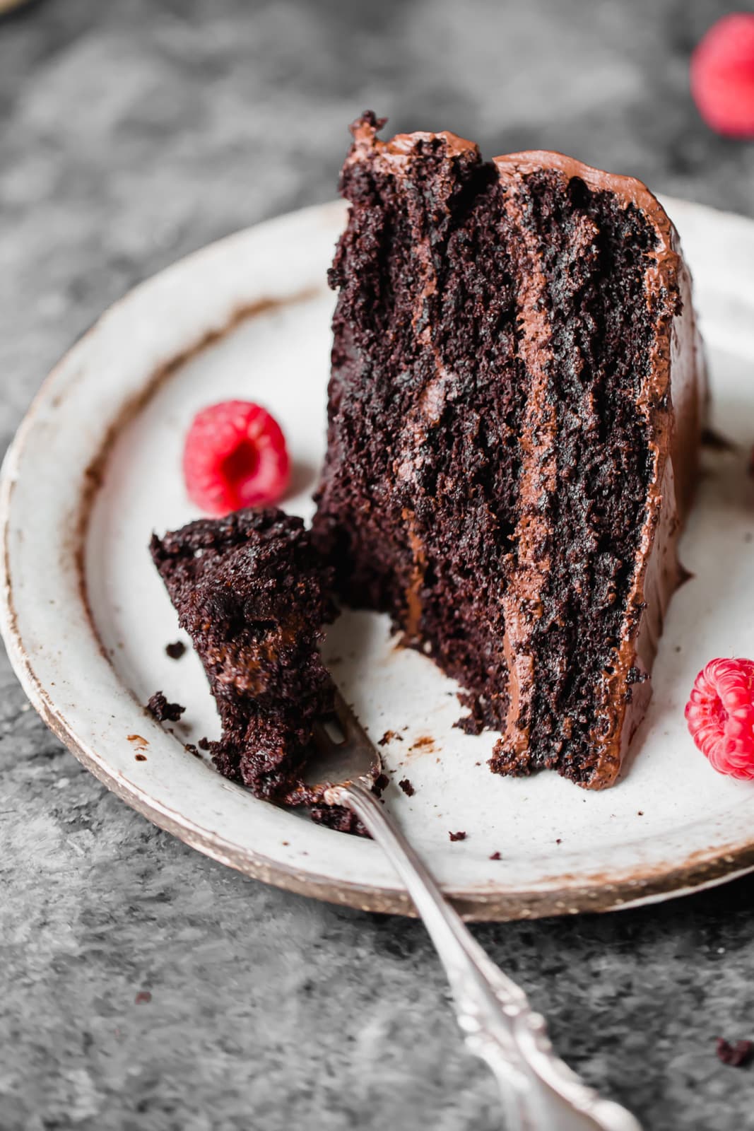 A slice of chocolate cake with chocolate frosting sits on a plate with raspberries; a fork holds a piece of the cake.