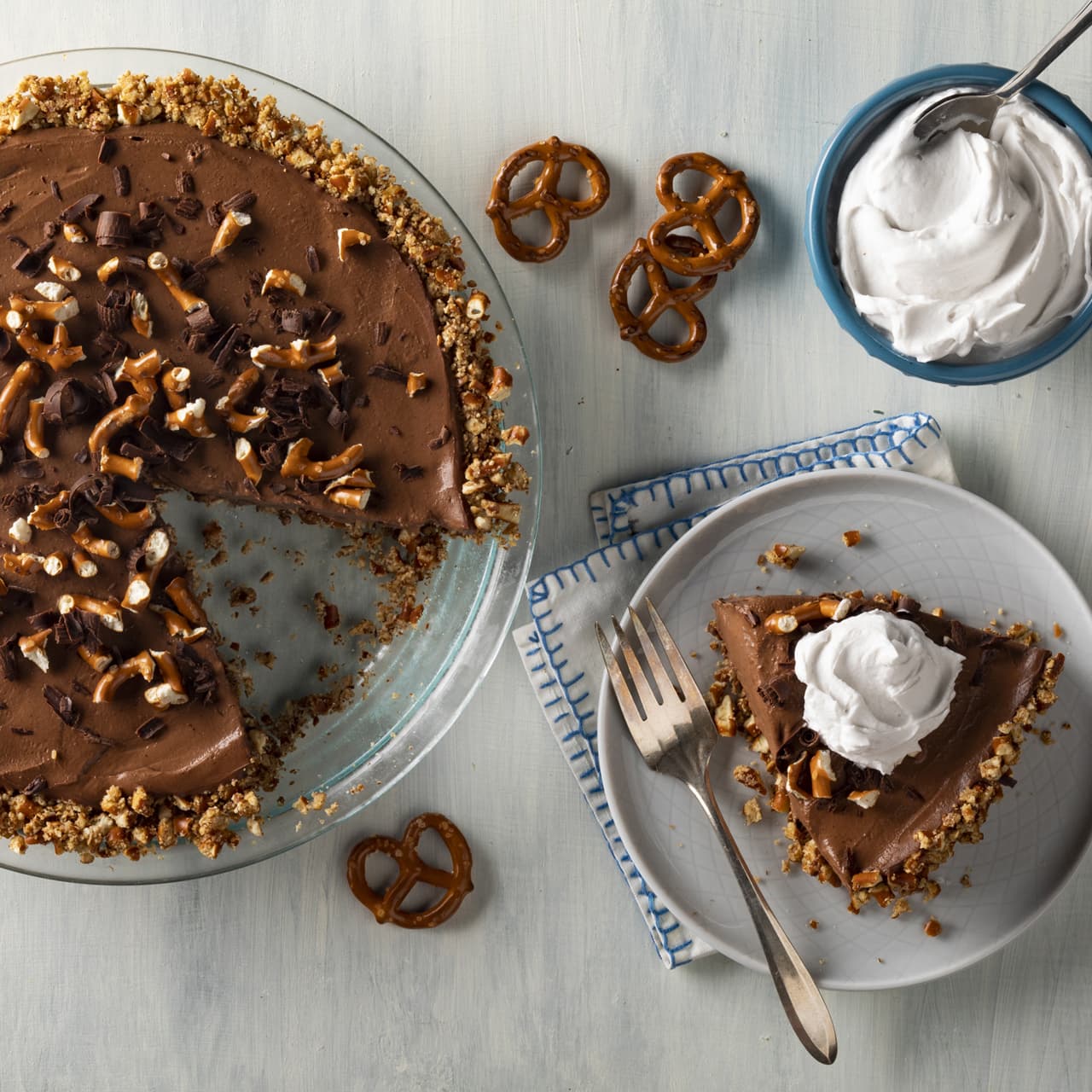 A chocolate pie with pretzel crust, topped with chocolate shavings and pretzels. One slice is served on a plate with whipped cream, next to a fork and a bowl of extra whipped cream.
