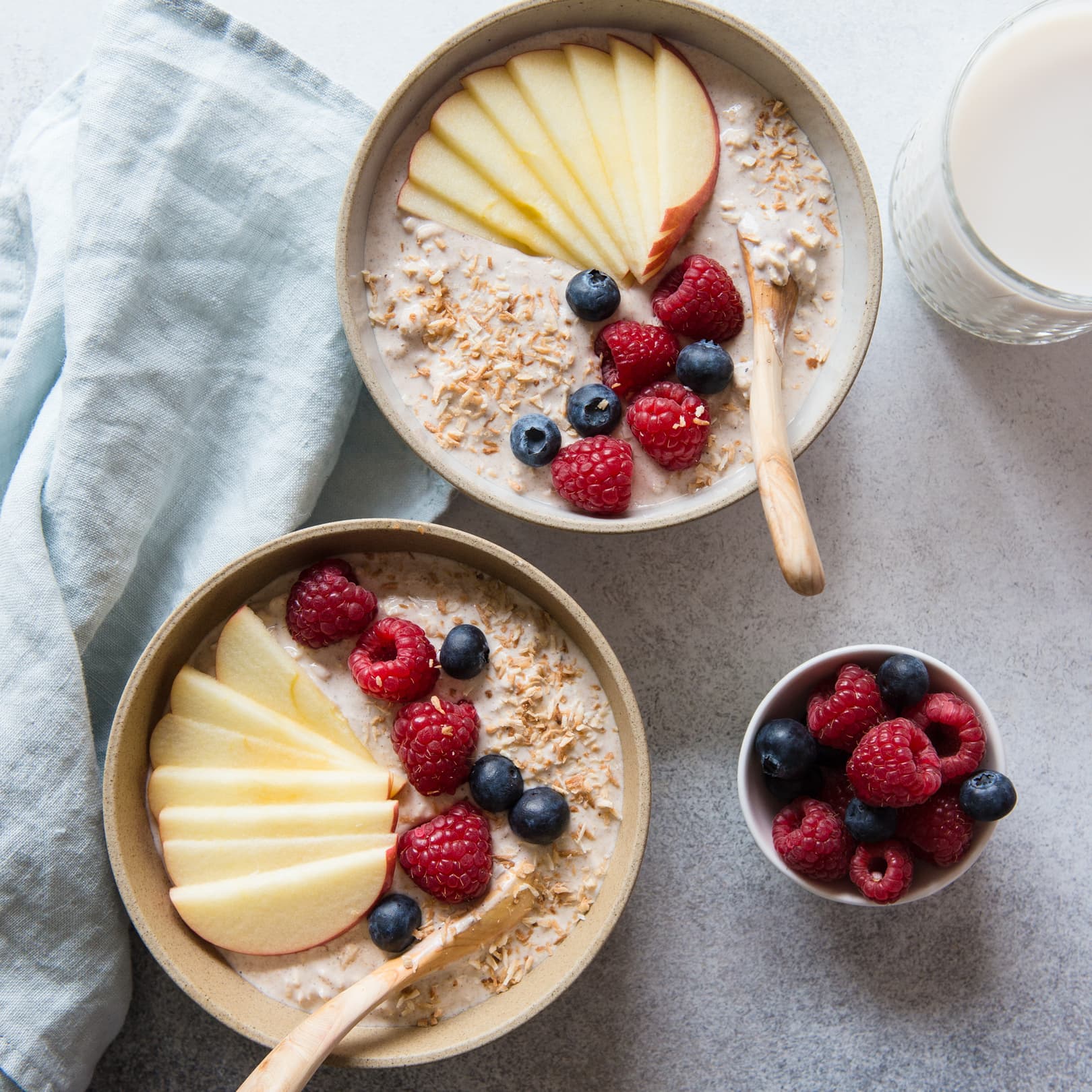 Two bowls of oatmeal topped with apple slices, raspberries, blueberries, and shredded coconut, next to a small bowl of berries and a glass of milk on a light surface.