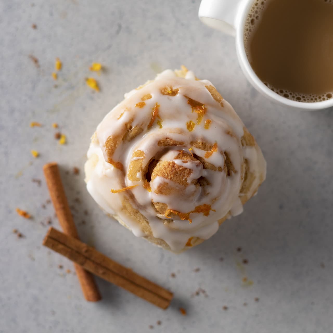 Iced cinnamon roll topped with orange zest next to two cinnamon sticks and a cup of coffee on a light gray surface.