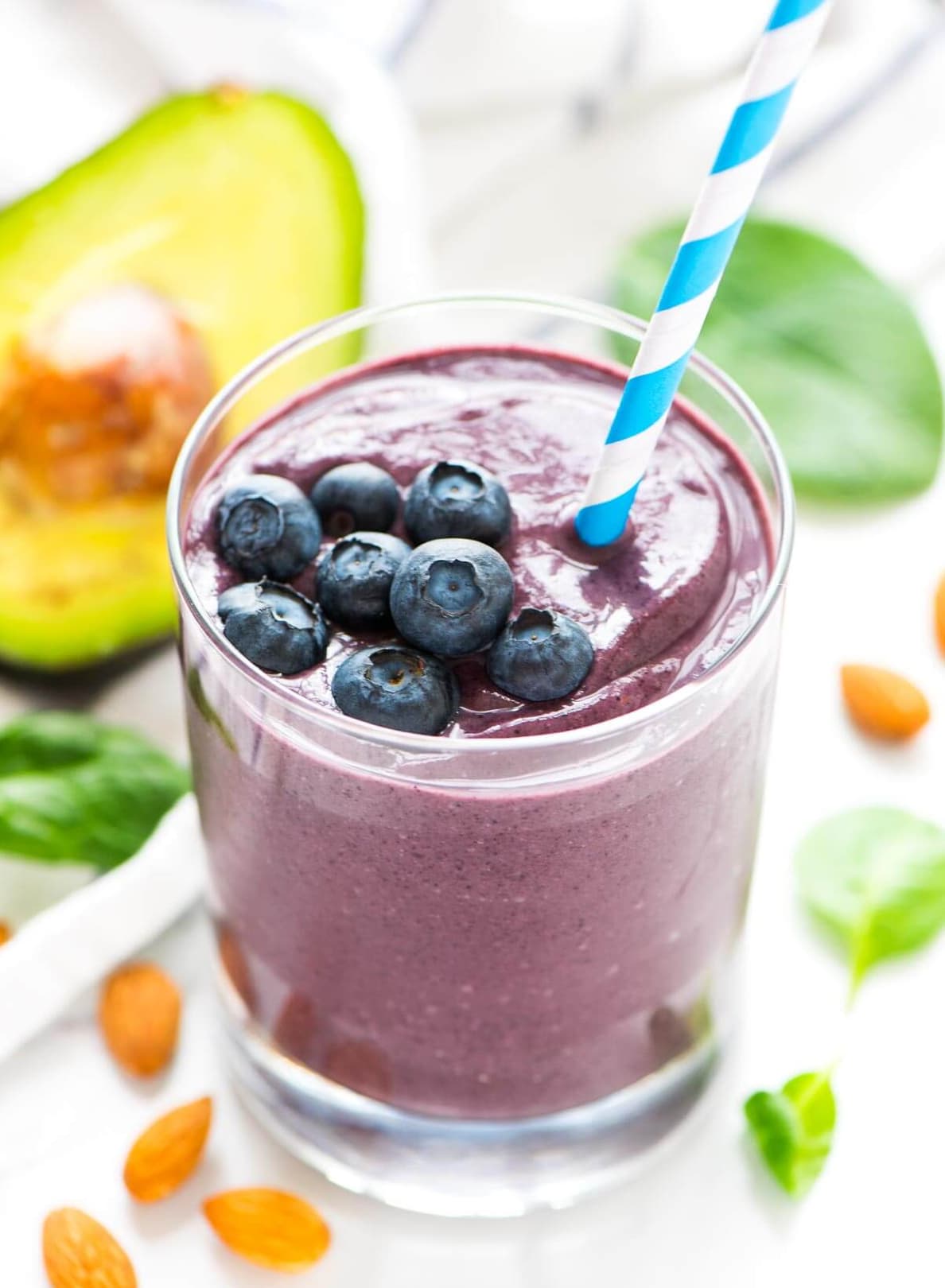 A glass of purple smoothie topped with fresh blueberries and a striped straw, surrounded by almonds, spinach leaves, and half an avocado.