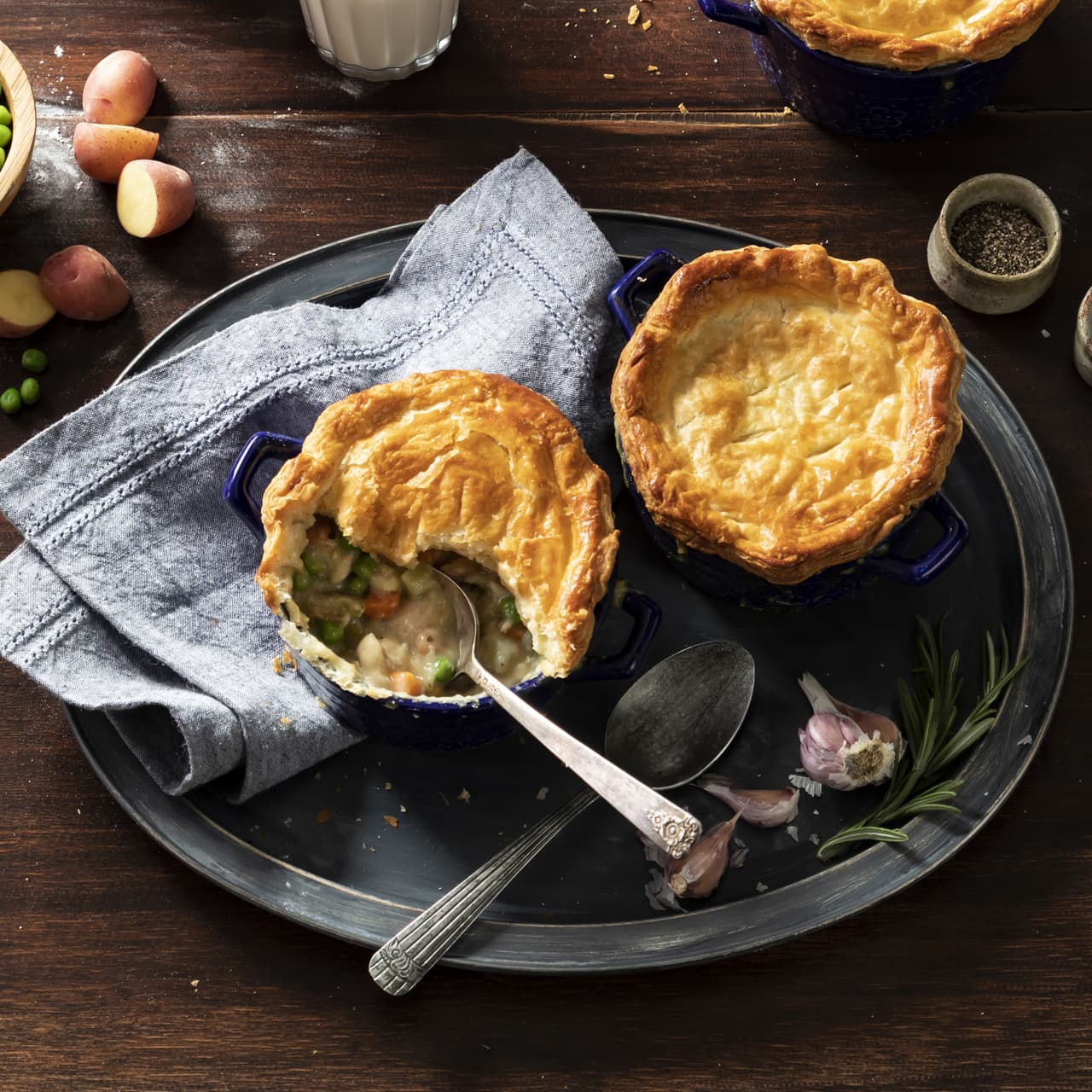 Two mini pot pies with golden crusts on a tray; one pie has a spoon and a portion missing, revealing a creamy filling with vegetables. Garlic, rosemary, and a napkin are nearby.