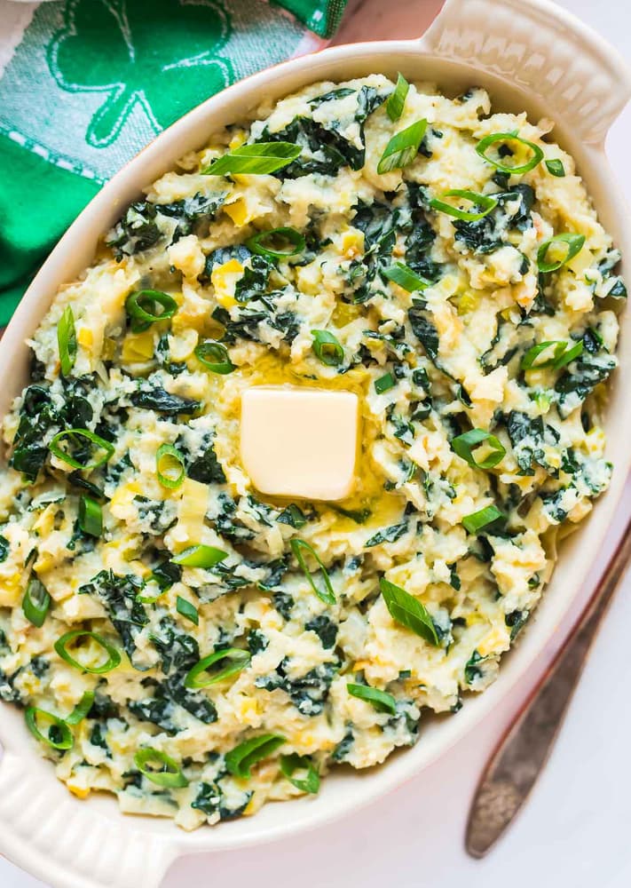 Oval dish of mashed potatoes mixed with chopped greens, topped with sliced green onions and a pat of butter, with a green napkin beside it.