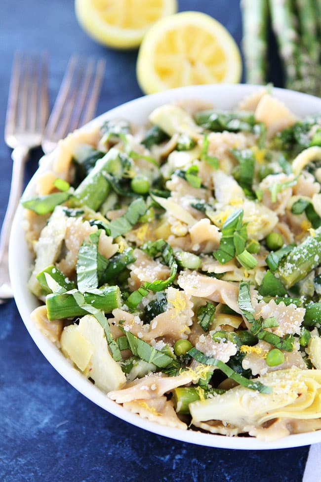 Bowl of farfalle pasta with green vegetables, artichoke hearts, grated cheese, and fresh herbs, placed on a dark surface with lemon halves and asparagus in the background.