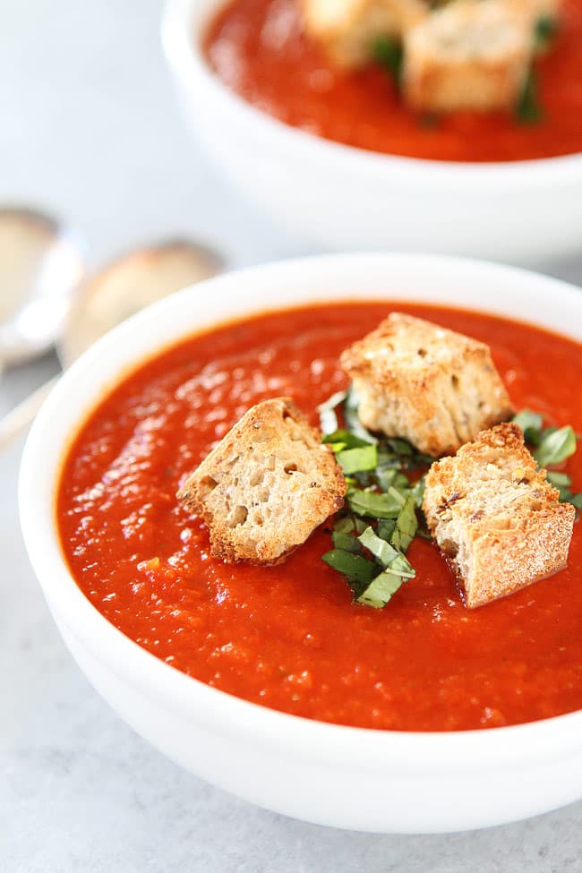 A white bowl of tomato soup topped with croutons and chopped basil, with another bowl and spoons blurred in the background.