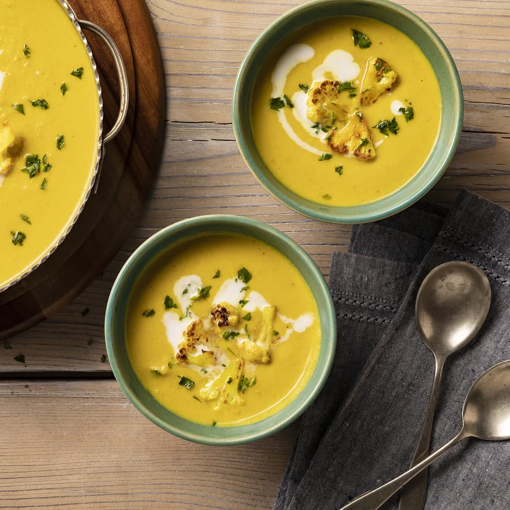 Two bowls of creamy yellow soup topped with roasted cauliflower, parsley, and a swirl of cream sit on a wooden table next to two spoons and a dark cloth napkin.