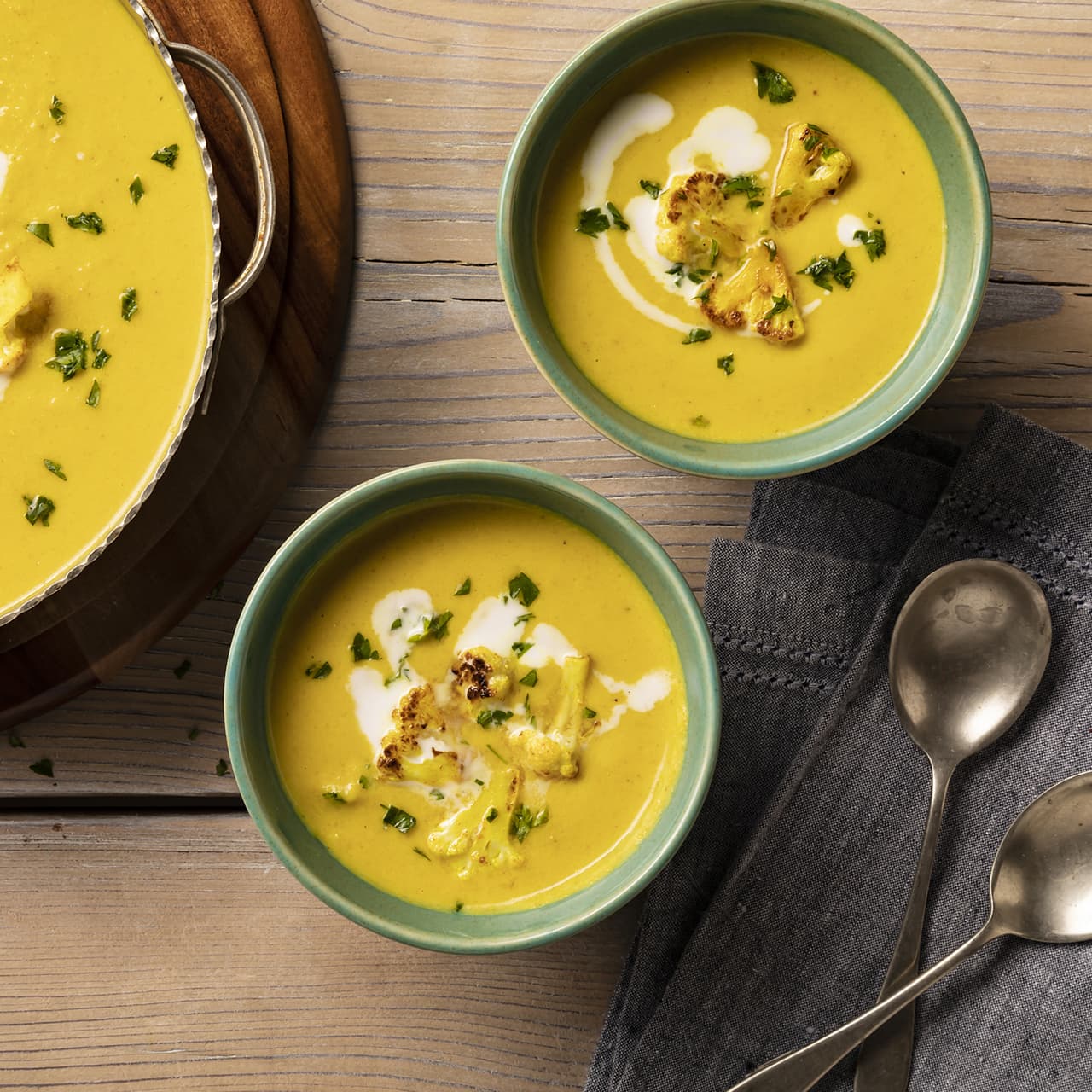 Two bowls of creamy yellow soup topped with roasted cauliflower, parsley, and a swirl of cream sit on a wooden table next to two spoons and a dark cloth napkin.