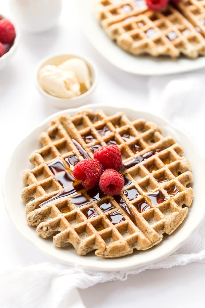 A waffle topped with syrup and raspberries is served on a white plate, with a small dish of butter in the background.