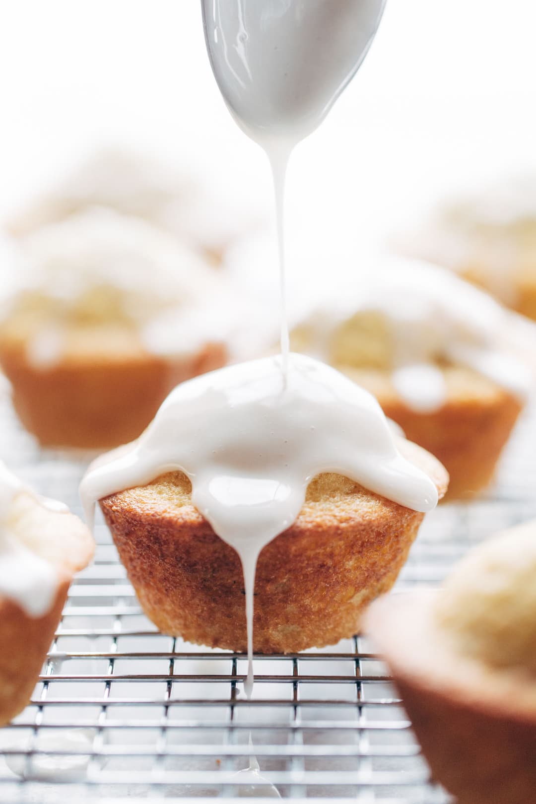 A spoon drizzles white icing onto a muffin resting on a wire cooling rack, with other muffins visible in the background.