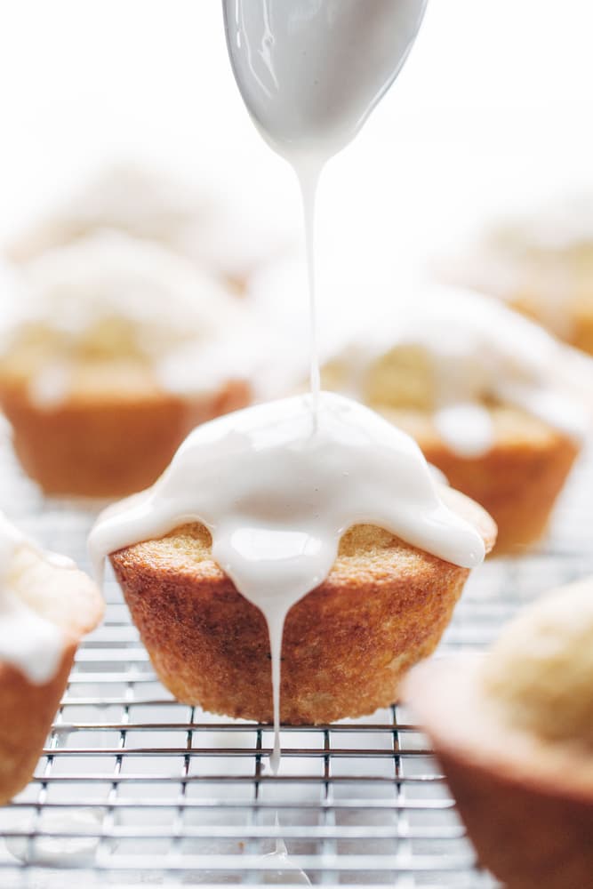A spoon drizzles white icing onto a muffin resting on a wire cooling rack, with other muffins visible in the background.