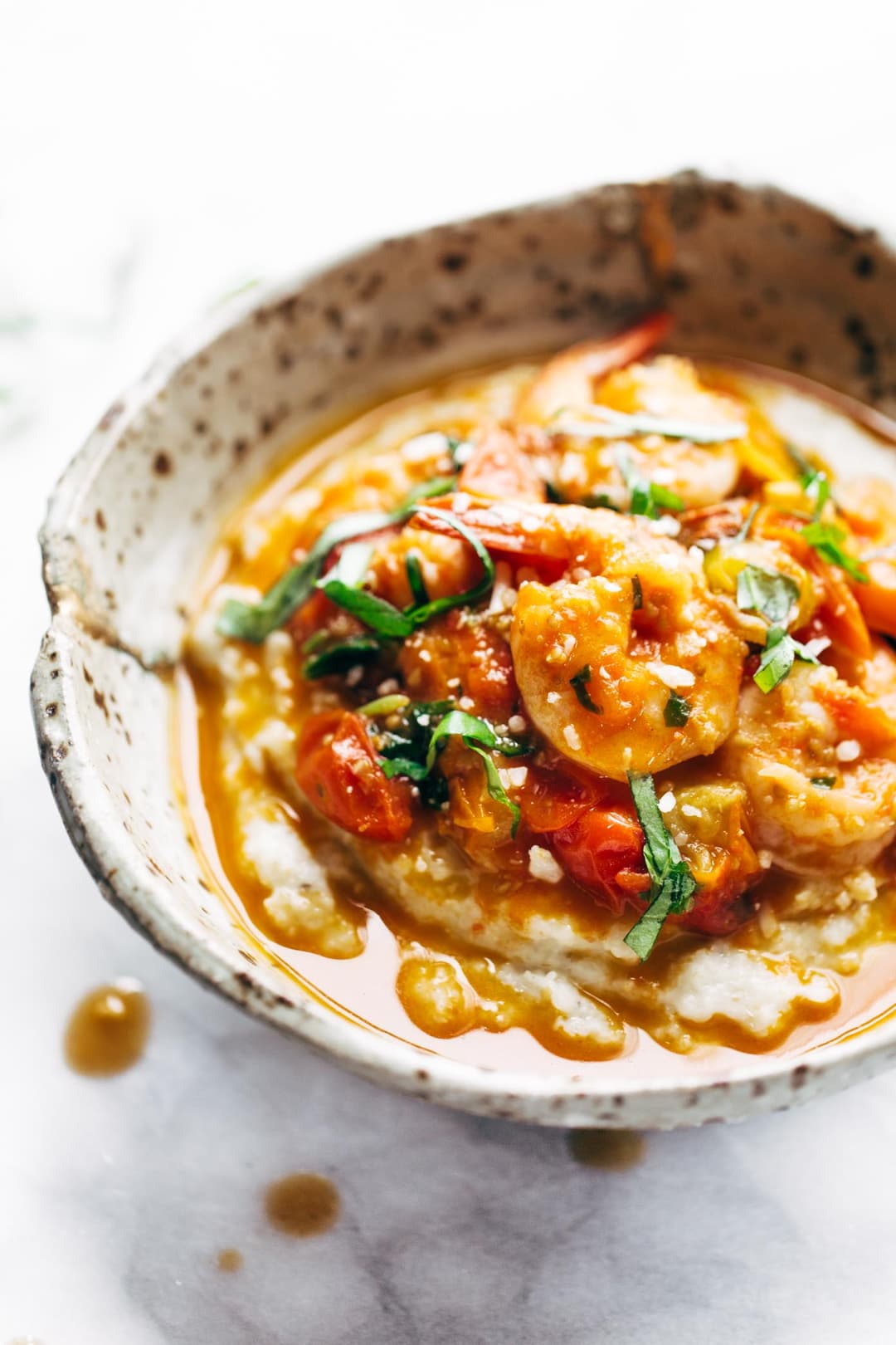 Bowl of shrimp and tomato stew served over grits, garnished with sliced basil, in a rustic ceramic dish on a marble surface.