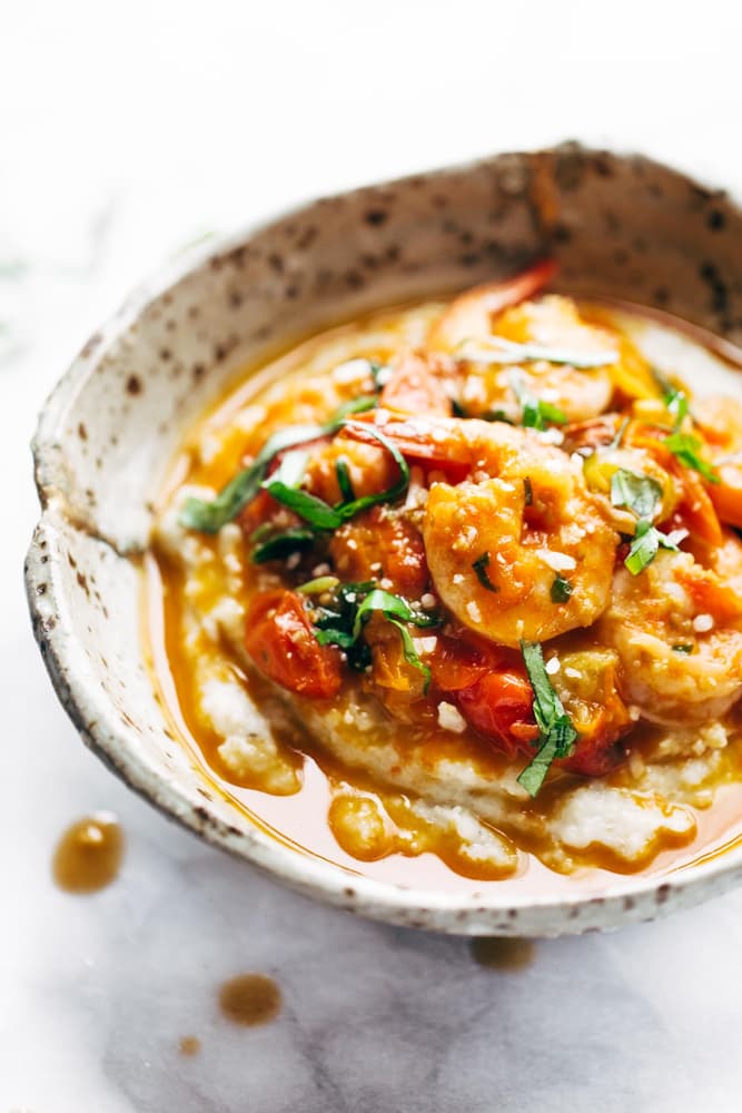 Bowl of shrimp and tomato stew served over grits, garnished with sliced basil, in a rustic ceramic dish on a marble surface.