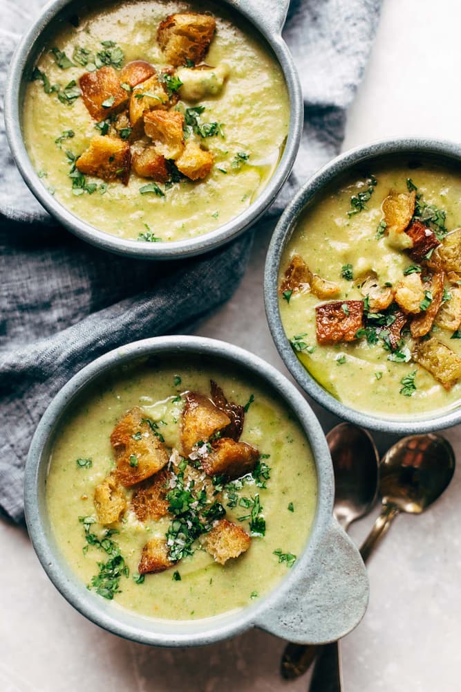 Three bowls of green soup topped with croutons and chopped herbs are placed on a gray cloth with two metal spoons beside them.