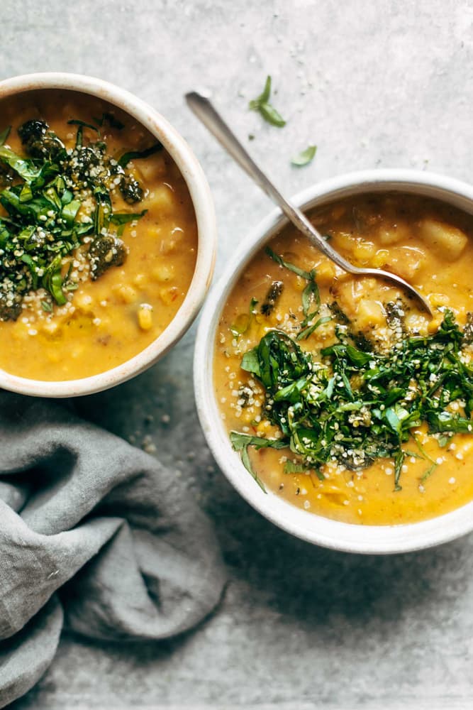 Two bowls of thick, yellow vegetable soup topped with chopped greens and seeds, with a spoon in one bowl and a gray cloth nearby on a light surface.