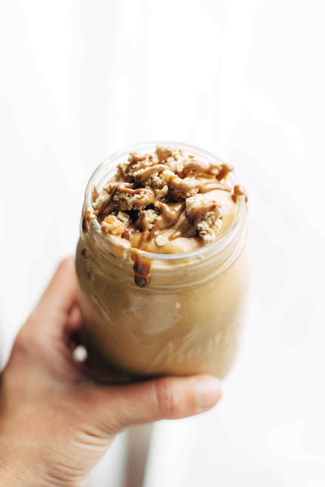 A hand holds a mason jar filled with a creamy smoothie, topped with granola and a drizzle of chocolate or nut butter, against a plain white background.