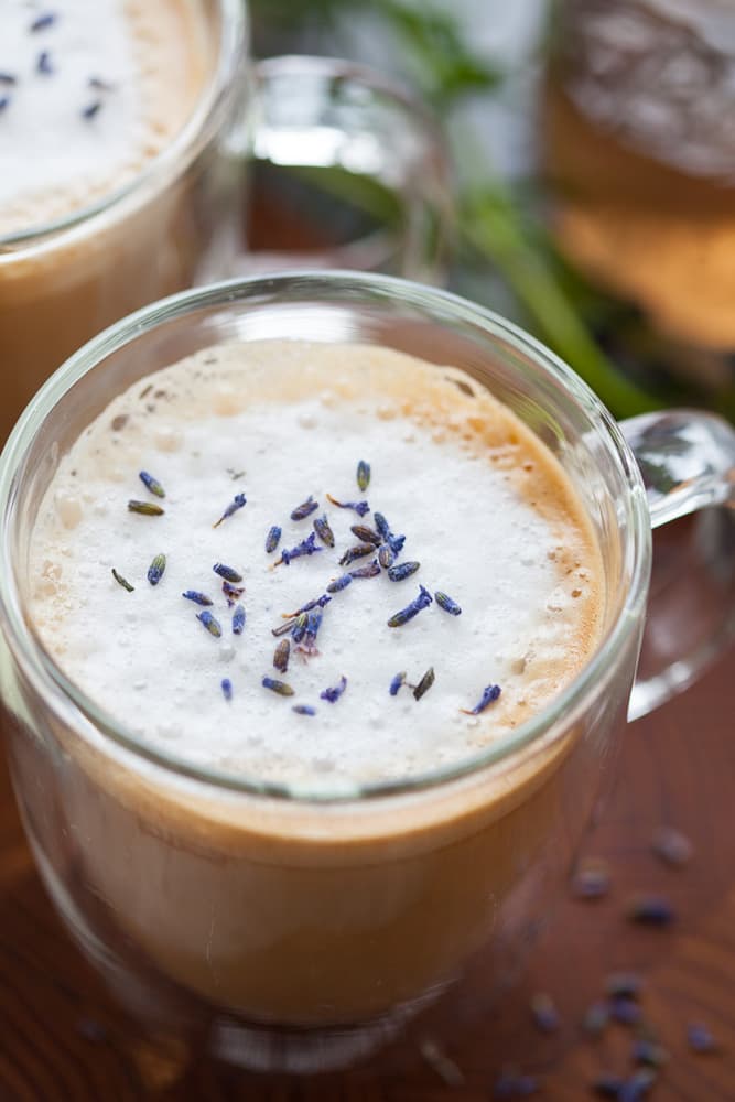 A glass mug filled with a foamy latte topped with dried lavender buds, placed on a wooden surface.