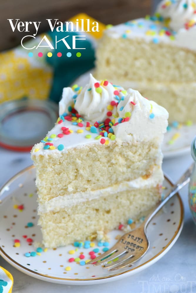 A slice of vanilla layer cake with white frosting and colorful sprinkles sits on a white plate with a fork.
