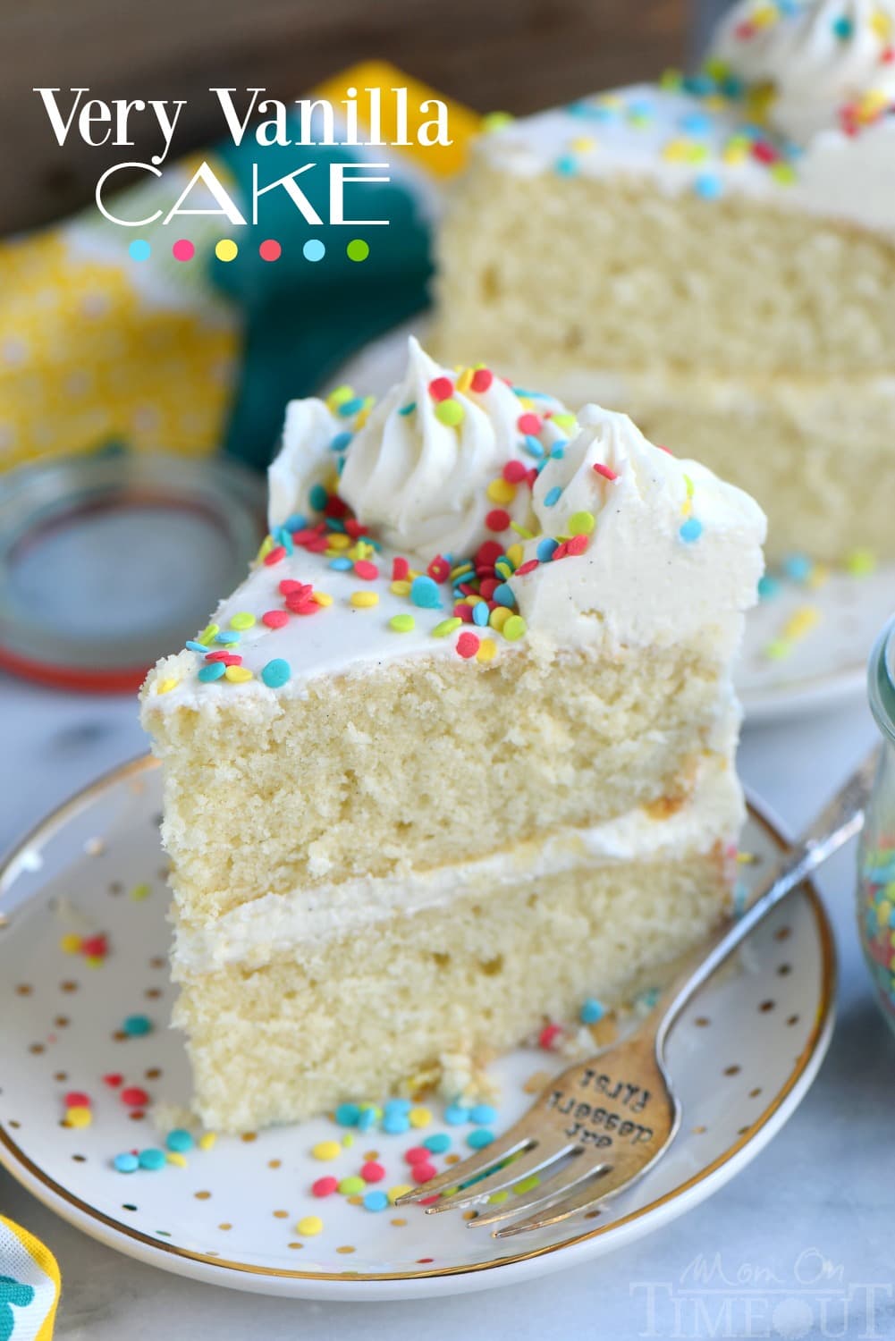 A slice of vanilla layer cake with white frosting and colorful sprinkles sits on a white plate with a fork.