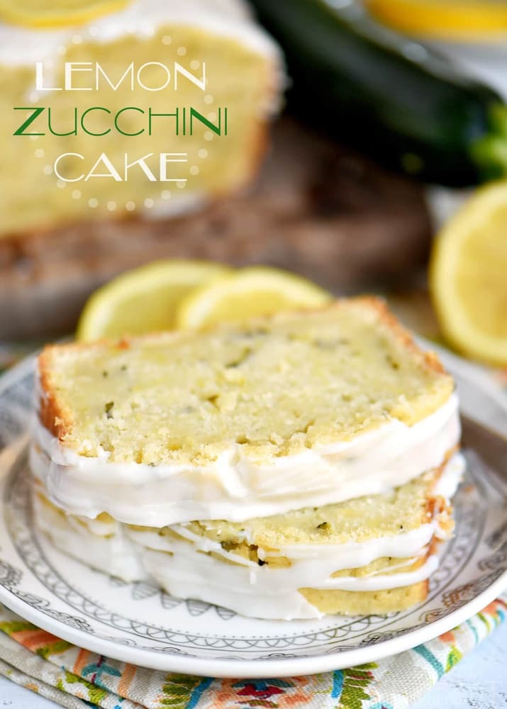 Two slices of lemon zucchini cake with white icing on a decorative plate, with lemon and zucchini in the background.