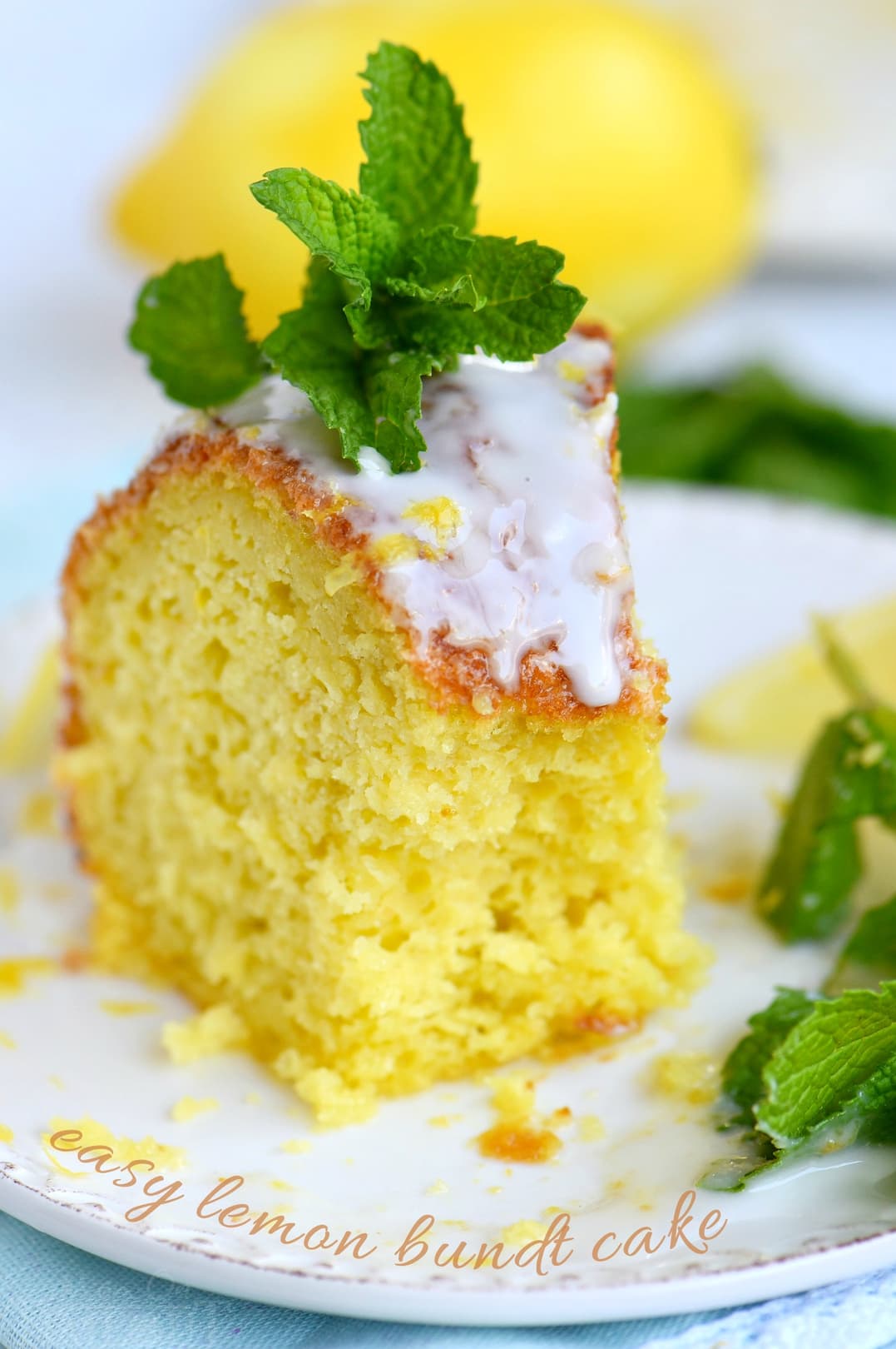A slice of lemon bundt cake with white glaze and a mint garnish is served on a plate, with a lemon and more mint leaves in the background.
