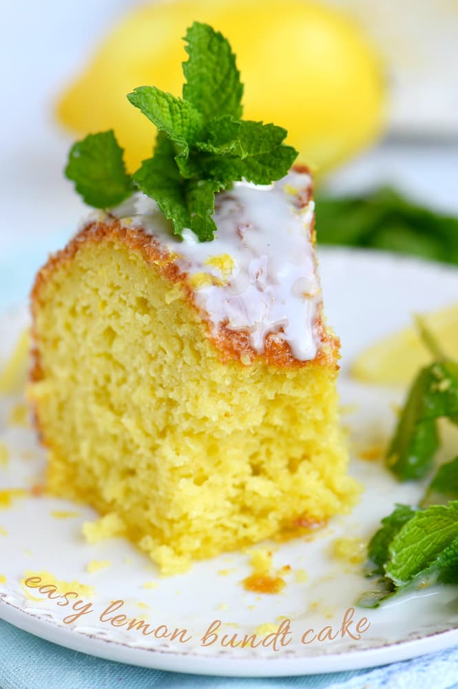A slice of lemon bundt cake with white glaze and a mint garnish is served on a plate, with a lemon and more mint leaves in the background.