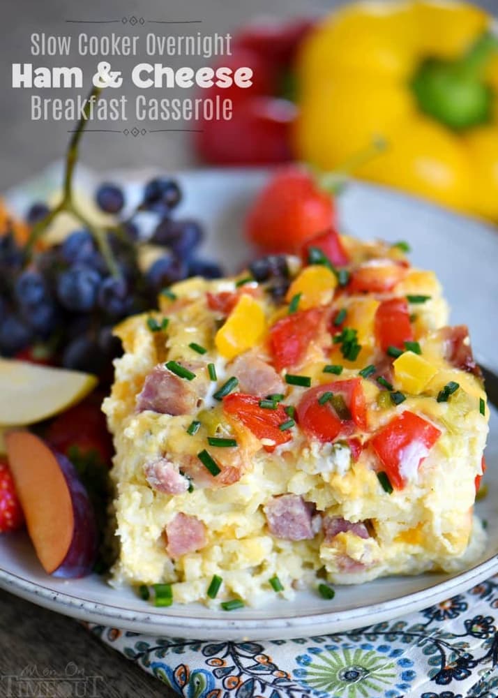 A slice of ham and cheese breakfast casserole garnished with chives, served on a plate with grapes, apple slices, and a strawberry.