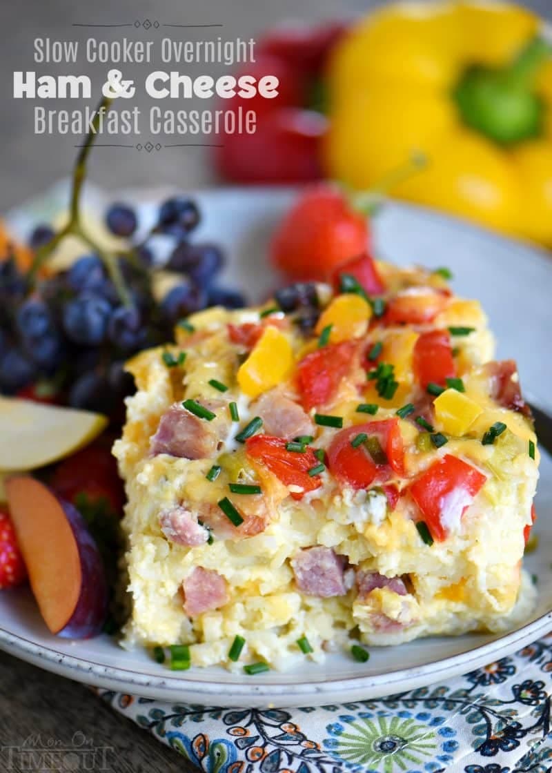 A slice of ham and cheese breakfast casserole garnished with chives, served on a plate with grapes, apple slices, and a strawberry.